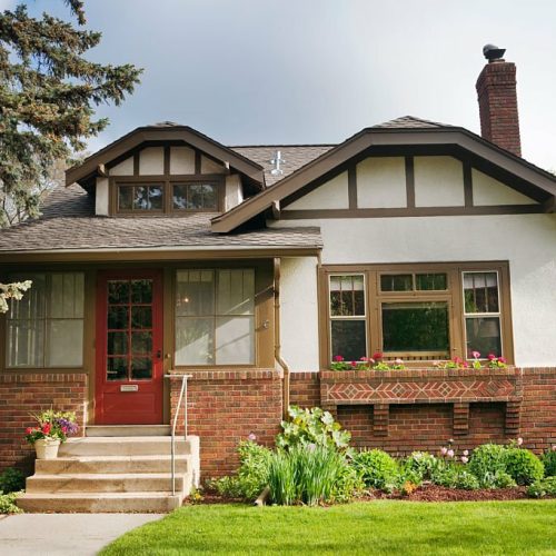 Full exterior front facade of 1920s era Arts and Crafts bungalow house. An example of Midwest, USA residential building style, the old, quaint architecture features stucco and brick walls, red front door, and picture window looking out to the lawn. A springtime flower garden grows outdoors. Horizontal, straight-on front view.